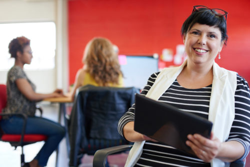 Casual portrait of a business woman using technology in a bright and sunny startup with the team in the background